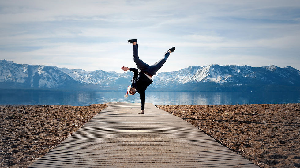 Person doing a handstand on a wooden path by a lake, wearing a Santa hat. Snowy mountains and blue sky in the background. Energetic mood.