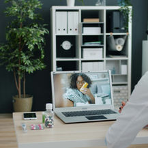 Doctor in white coat watches a video call with a patient holding a yellow inhaler in a neat office with shelves and plants.