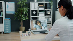Doctor in white coat watches a video call with a patient holding a yellow inhaler in a neat office with shelves and plants.