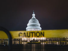 US Capitol at night illuminated, behind yellow caution tape. Dark sky backdrop, creating a tense atmosphere.