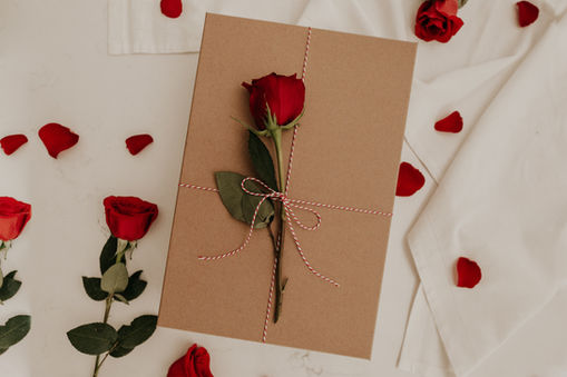 Brown gift box with a red rose and striped twine on top, surrounded by scattered red rose petals on a white surface, creating a romantic mood.