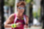 Female runner in pink tri suit and reflective sunglasses jogging outdoors, focused expression, blurred trees and walkway behind her