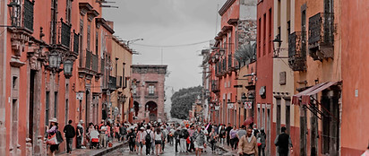 A bustling street in a historic city with colorful colonial buildings, balconies, and many people walking along the cobblestone road under a cloudy sky.