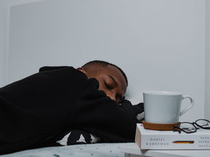 Person sleeping on bed, wearing a black hoodie. Nearby are a white mug, glasses, and two books.