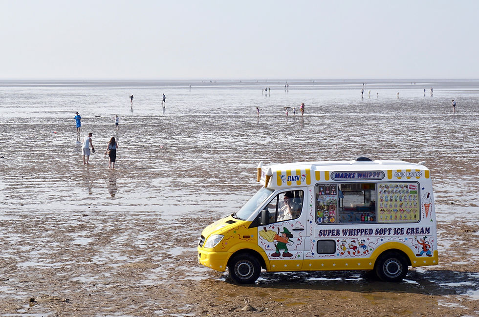 A yellow and white ice-cream van, on a flat, wet beach. A sparse amount of people play in the sand. The weather is overcast.