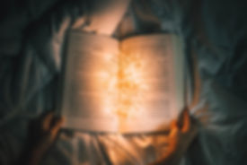 A pair of hands hold open a bookon a bed cover, with fairy lights in the centre. Image courtesy of Nong Vang via Unsplash.