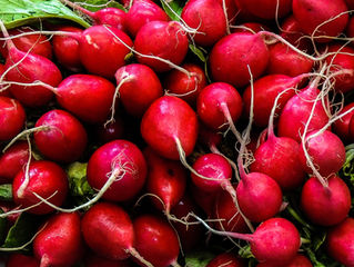 A pile of vibrant red radishes with green leaves, displaying thin roots. The fresh produce is closely packed, showcasing a natural pattern.
