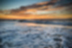 Ocean waves with frothy surf under a colorful sunset sky. Orange, blue, and gray hues create a peaceful, serene atmosphere.