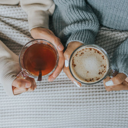 Two hands hold mugs on a cozy knitted blanket. One mug has tea, the other has frothy coffee. Warm, relaxed atmosphere with neutral tones.