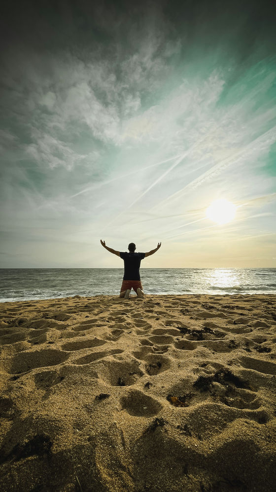 Embracing the tranquility of the ocean, a person kneels on the sandy shore, arms outstretched in gratitude as the sun sets on the horizon.