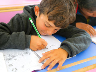 Young boy in a classroom writes in a notebook with a green pencil, focused, while another student works beside him.
