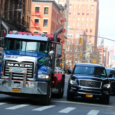 Blue Mack truck and black SUV drive closely on a city street with tall buildings. Bright cones and road signs in the background.
