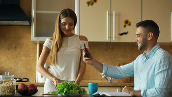 Woman chopping greens in a kitchen. Man smiles, holding phone. Background shows wooden cabinets with sunflower decor. Relaxed mood.