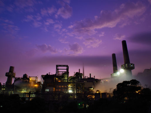Industrial plant with tall chimneys emits smoke under a purple sky at dusk. Bright lights illuminate the complex, creating a moody scene.