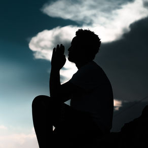the silhouette of a man praying, sitting on a rock with a moody sunset background.