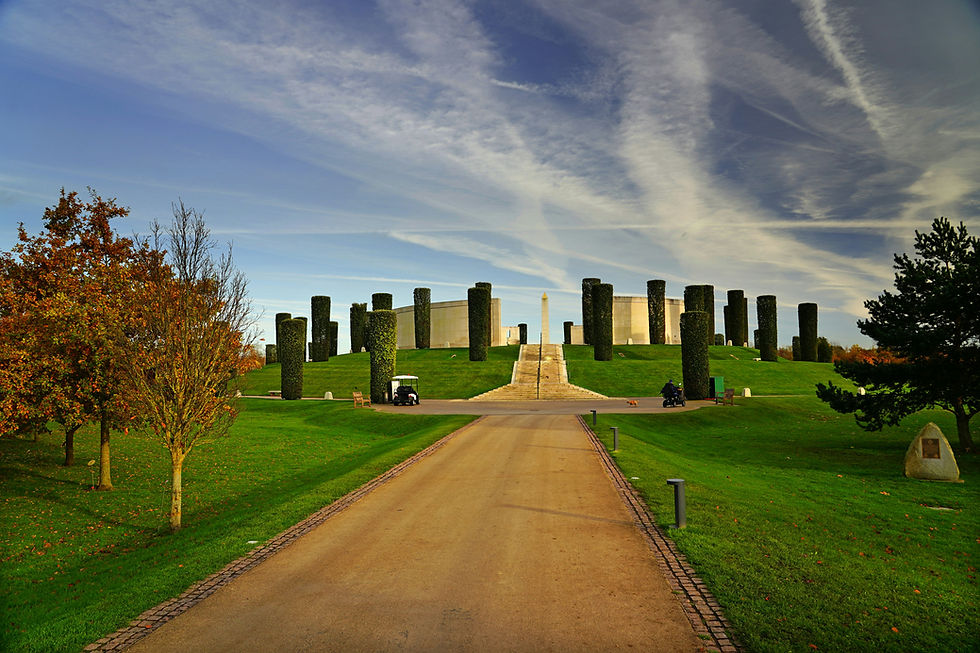 Landscaped memorial hill with an obelisk, tall clipped hedges, a central path, autumn trees, and a dramatic cloudy sky.