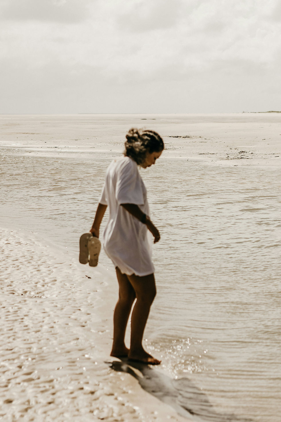 Woman in white dress strolls barefoot on a sandy beach, holding flip-flops. Calm water and cloudy sky create a serene atmosphere.