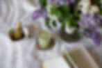 A matcha latte with a bamboo whisk and book on a white cloth, next to a vase of purple and white lilacs, creating a calm, cozy scene.