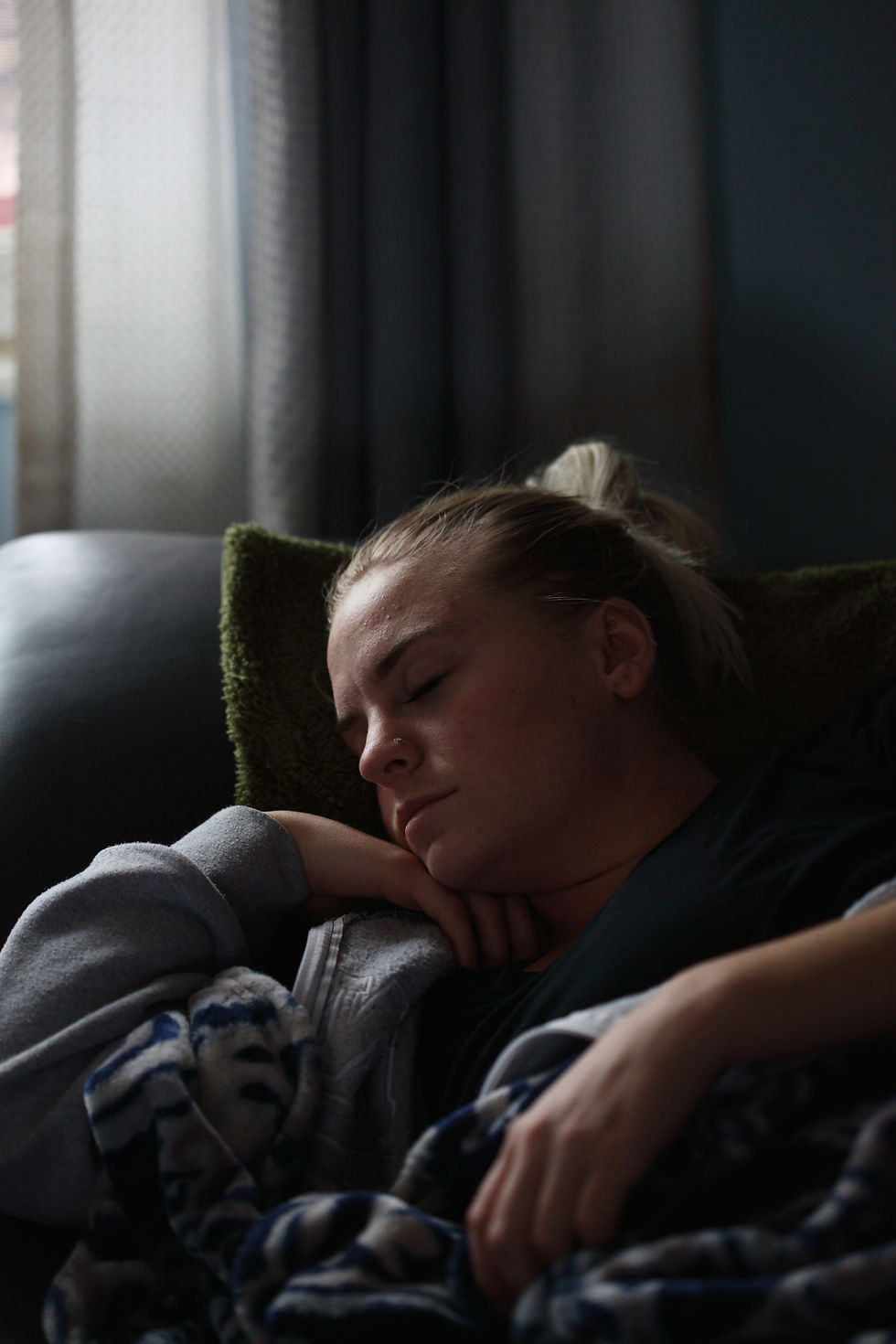 Person sleeping on a couch, wrapped in a blue patterned blanket. Soft lighting and dark curtains create a calm, cozy atmosphere.
