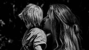 Woman holding a child in a tender embrace, both looking away in a dark, natural setting. Black and white with soft, contemplative mood.