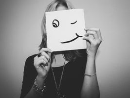 Person holding a square paper with a winking face drawn on it, covering their face. Black and white image with playful mood.