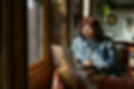Person on a sofa using a laptop beside a sunny window. Room has warm decor, plants, and shelves. Cozy, focused mood.