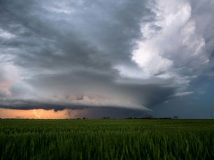 Massive supercell thunderstorm over a green field, with lightning under dark swirling clouds and an orange-lit horizon.