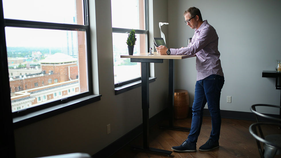 A man is standing by a standing desk by a window. There's a laptop on the table and he's looking at his phone. He's leaning on his left leg which is locked back. His right leg is forwards and not making much contact with the floor. His spine is slightly rotated to the left and his head is leaning down a bit
