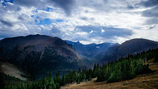Cloudy mountain valley with rugged peaks, evergreen forest, and golden grass under dramatic blue-gray sky