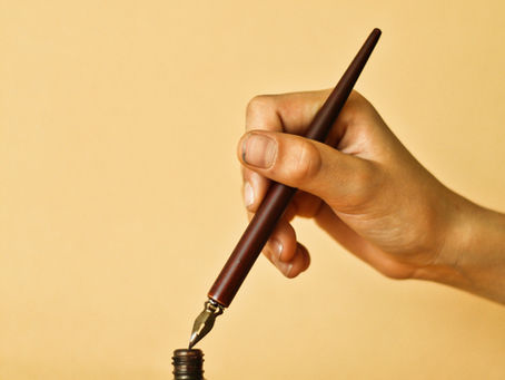 Hand dipping a fountain pen into an ink bottle on a gold stand; beige background, label reads Inchiostro Stilografico, Kaligrafica.