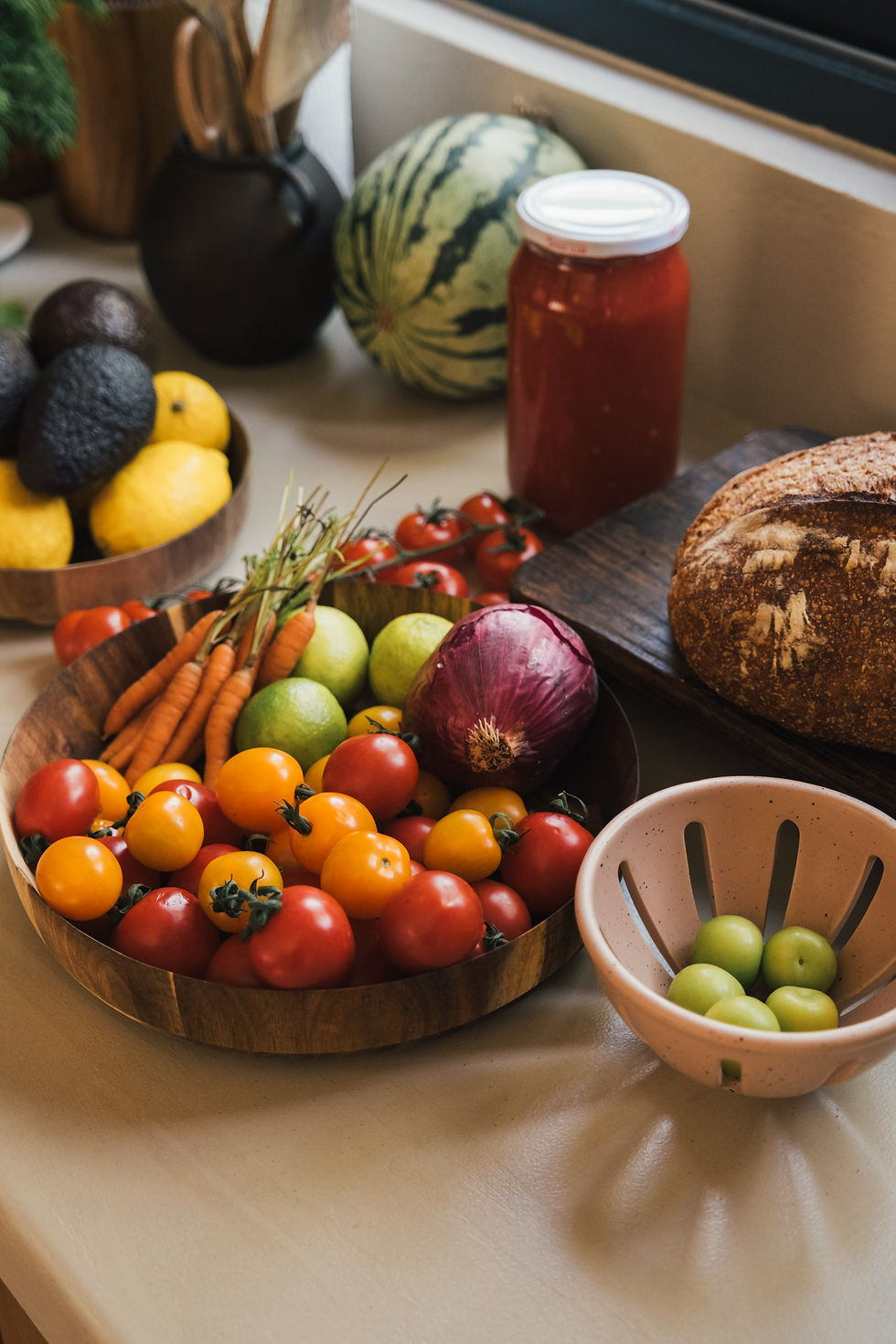 Colorful kitchen countertop with bowls of tomatoes, carrots, onions, avocados, citrus, bread, watermelon, and a jar of sauce