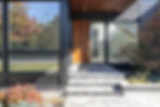 Modern house entrance with wooden door, glass panels, and gray stone steps. Reflections of trees in windows and greenery around.