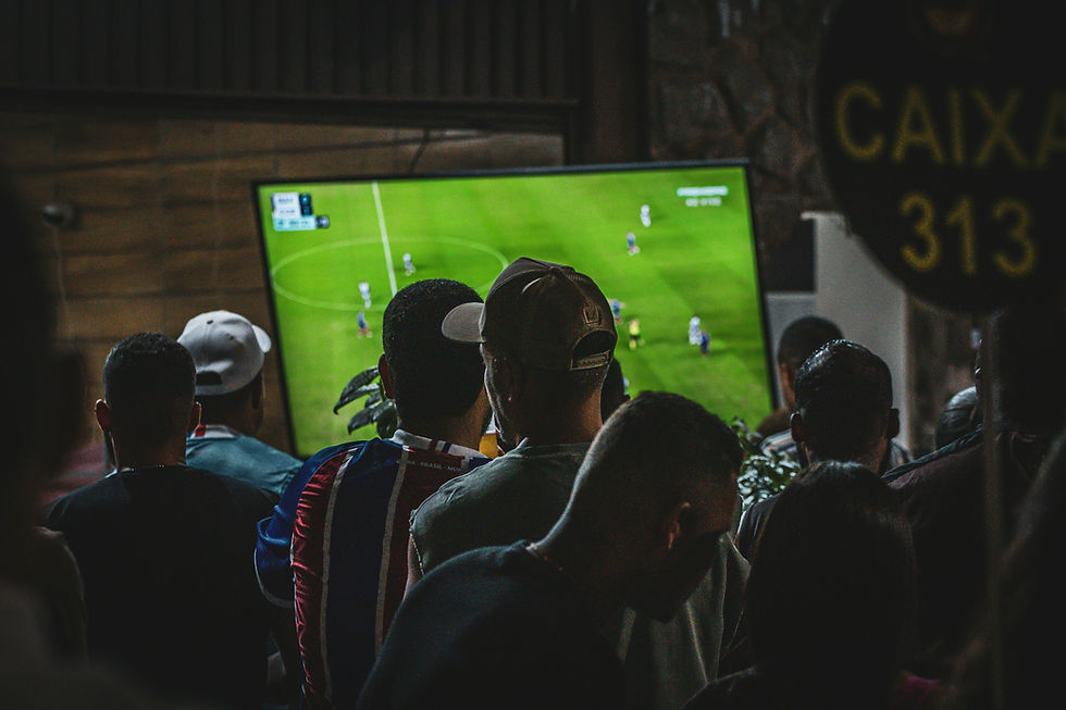 People watch a soccer match on a TV screen in a dimly lit room. The crowd is focused, and a "Caixa 313" sign is visible.