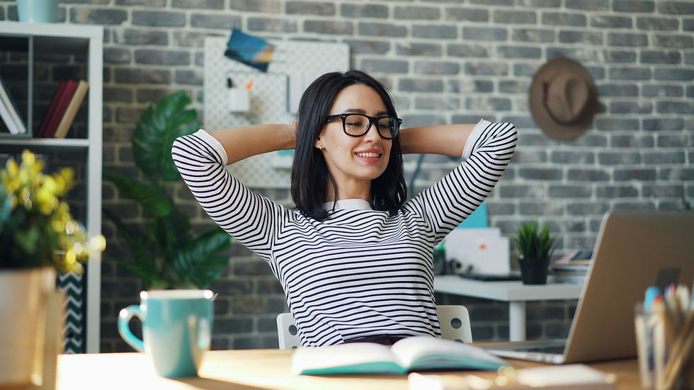 Smiling woman in striped shirt smiling at laptop. Hands behind her head and grey brick wall in background, bright setting.