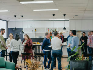 People chatting and socializing in a modern office break room. Casual attire, a few holding drinks. Plants and pendant lights decorate the space.
