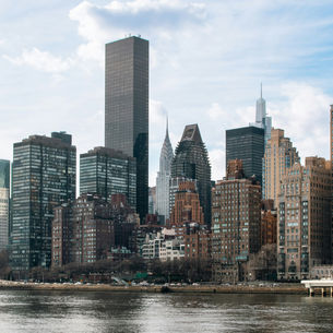 Midtown Manhattan skyline viewed from the East River, with waterfront apartment buildings and the Chrysler Building beneath a partly cloudy sky.