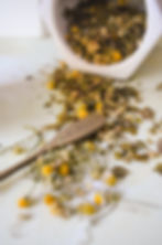 A rustic and heartfelt scene featuring dried chamomile and herbal tea ingredients spilling from a white container onto a light surface. A wooden spoon engraved with "made with love" rests among the herbs, adding a personal and nurturing touch. The warm tones and natural textures evoke a sense of care, tradition, and the mindful craft of herbal tea blending.