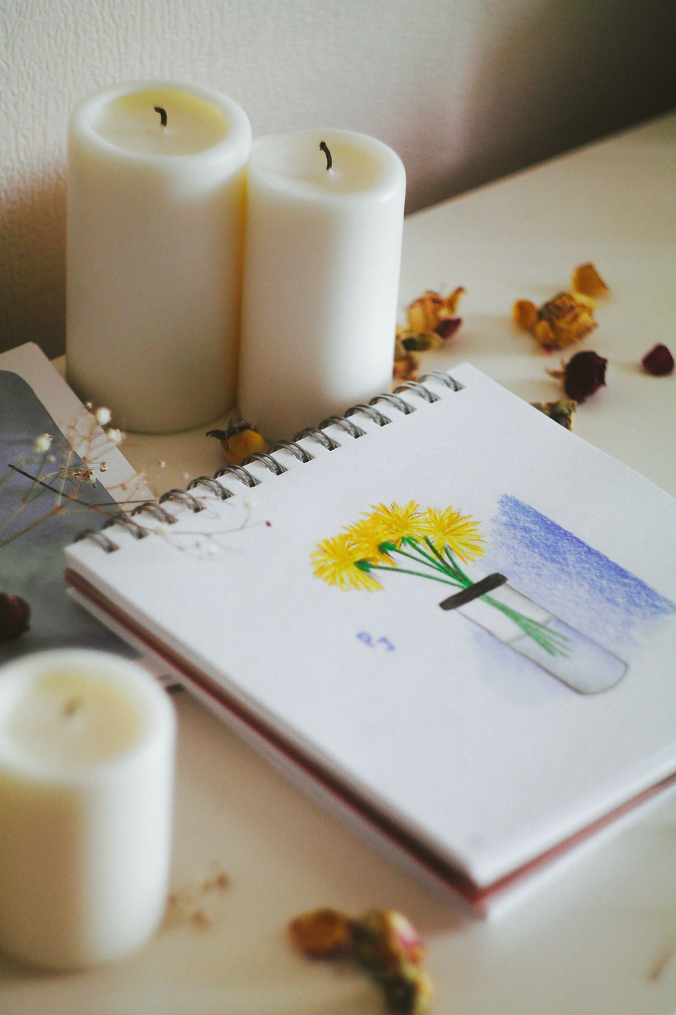 Two white candles beside a spiral sketchbook with a yellow flower drawing and dried petals on a soft, calm tabletop scene