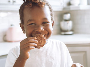 Happy young boy with curly hair, smiling while eating a gluten-free cookie at home.