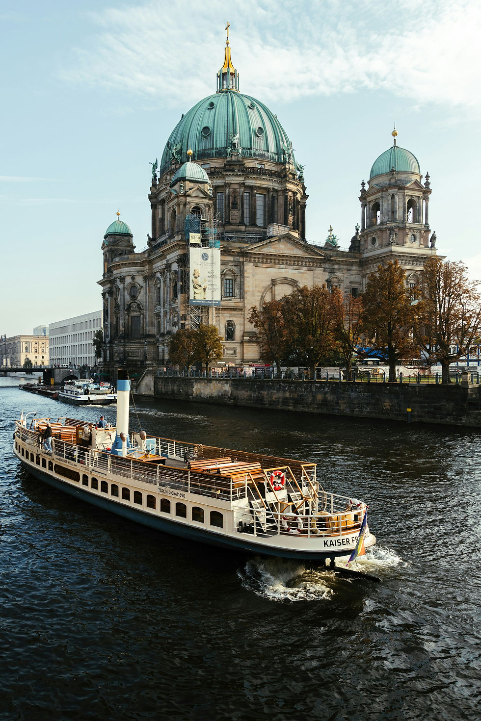 A historic boat cruises along the river, passing by the grand Berlin Cathedral on a crisp autumn day.