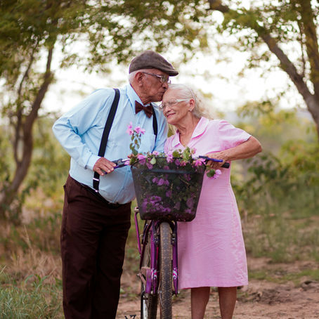 Elderly couple in a park, man kisses woman's forehead beside a purple bike with flowers. She wears pink, he wears blue. Peaceful setting.
