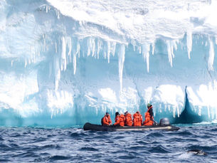 People in orange jackets on a boat near icy cliffs with icicles in Antarctica. Blue ocean waves, and a penguin swimming in foreground. Cold, adventurous scene.