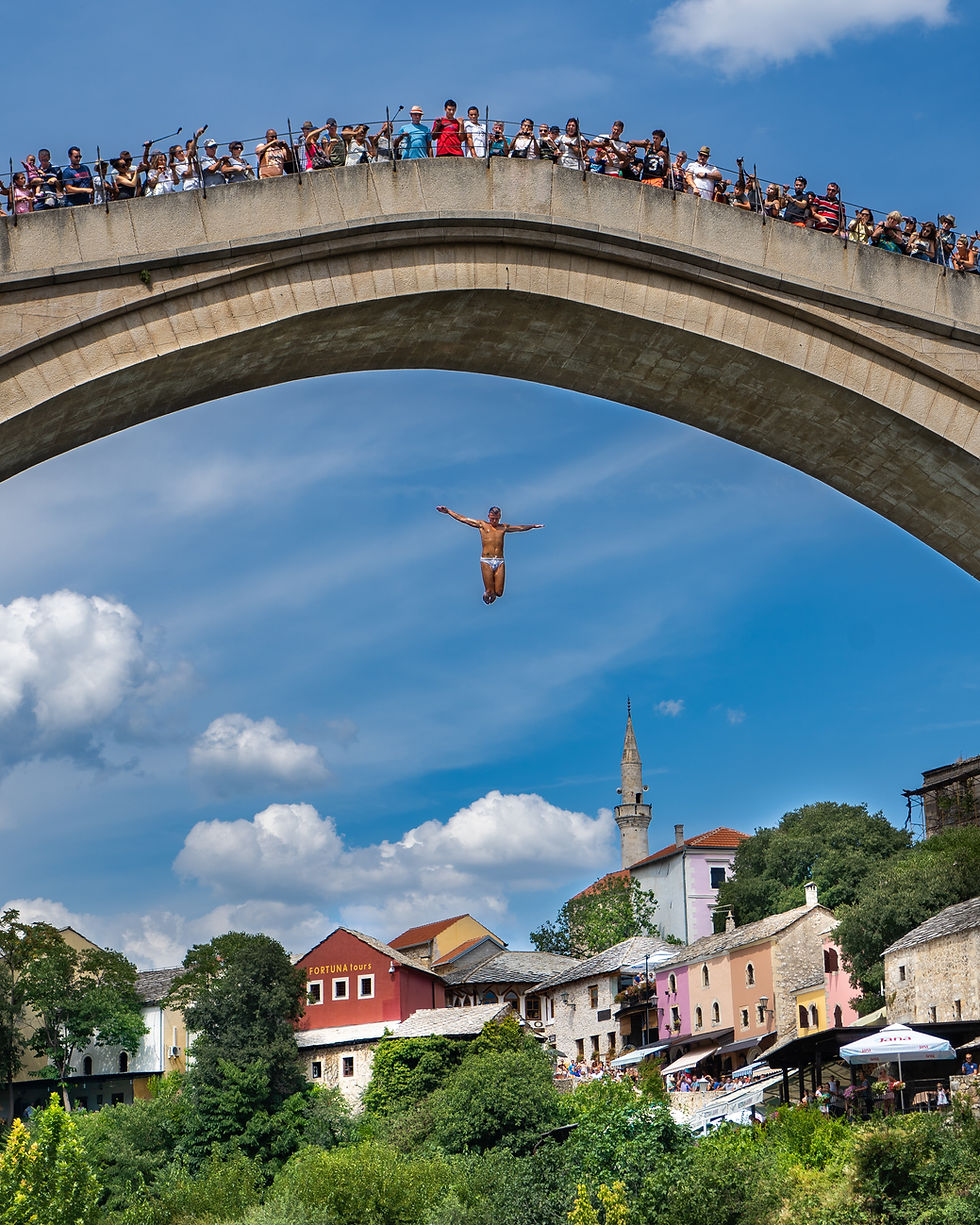 A diver jumps from a stone bridge into a river, arms outstretched. Crowd watches atop the bridge. Colorful buildings and mosque in background.