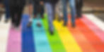 numerous feet cross a rainbow painted crosswalk. one of the people in the middle has a prosthetic leg.