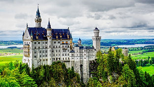 A large, fairy-tale castle with tall towers and spires sits atop a forested hill, overlooking a green valley and a distant lake under a cloudy sky.