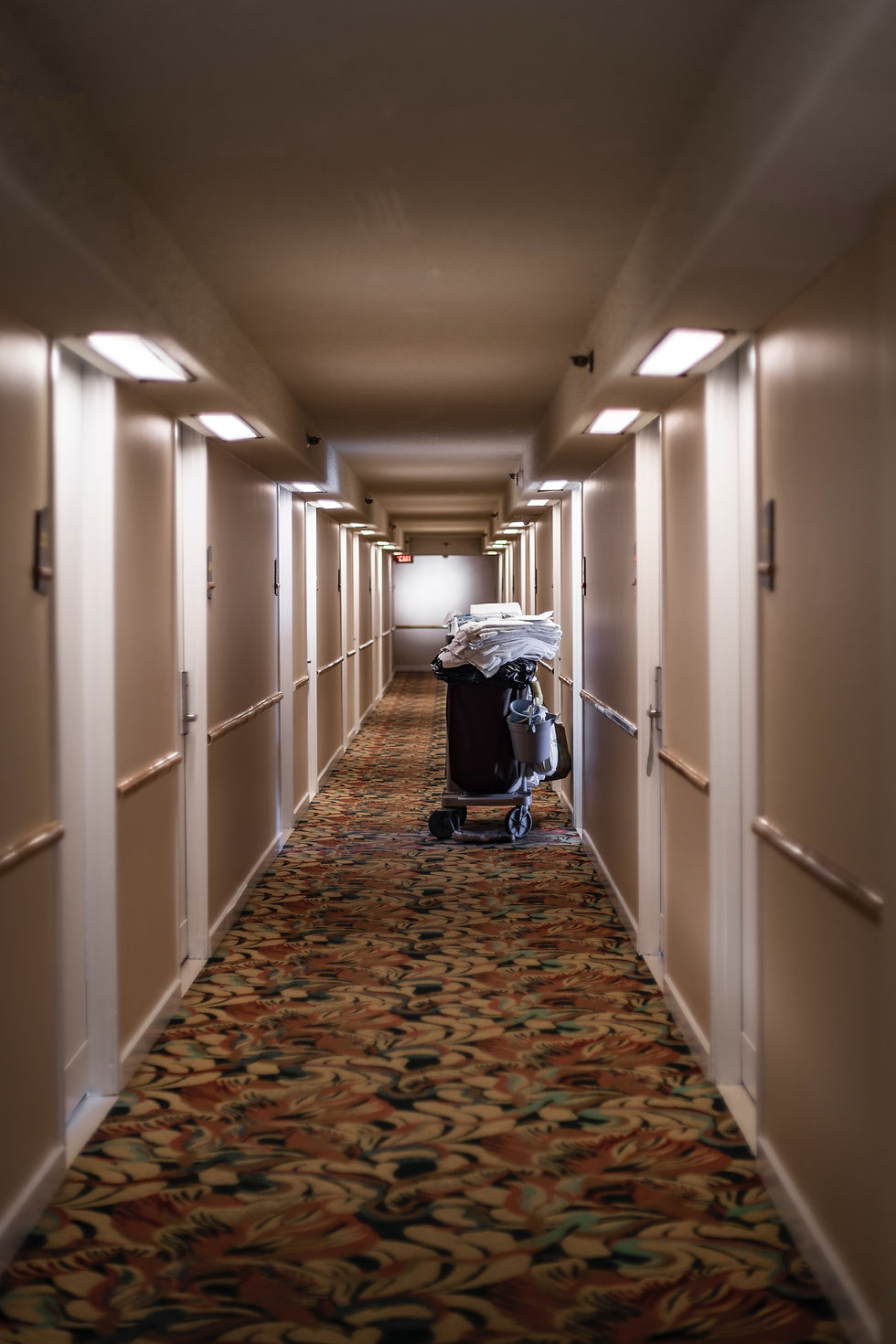 Empty hotel hallway with a housekeeping cart piled with white towels on patterned carpet under warm lights.