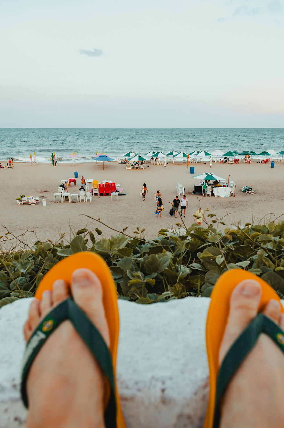 Feet in orange flip-flops overlook a calm beach with umbrellas, people, and the ocean at dusk.