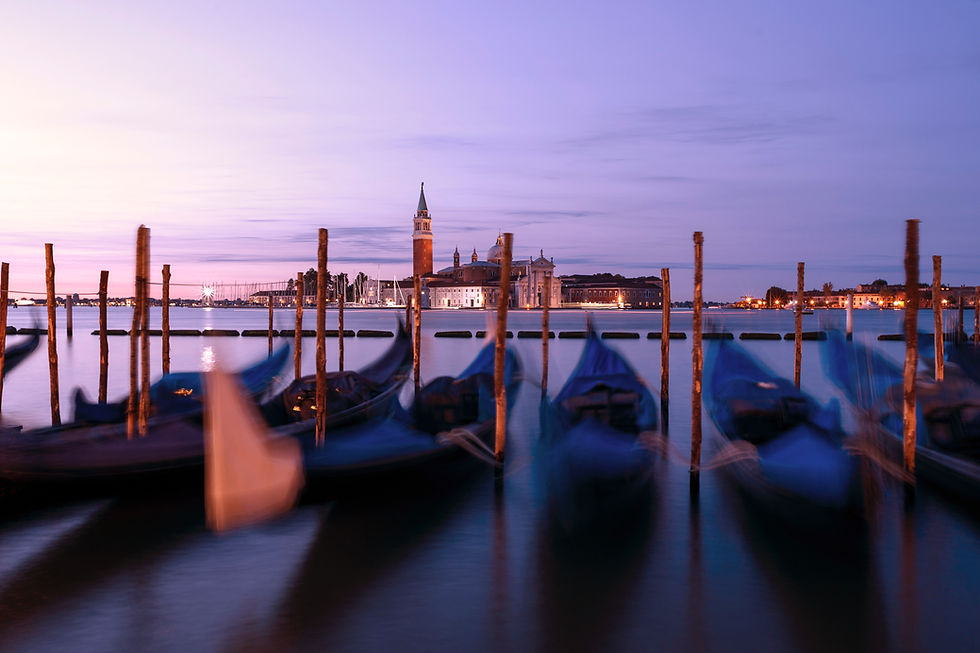 Blurred gondolas on calm water at dusk in Venice, with San Giorgio Maggiore in the background. Sky and water have a purple hue.