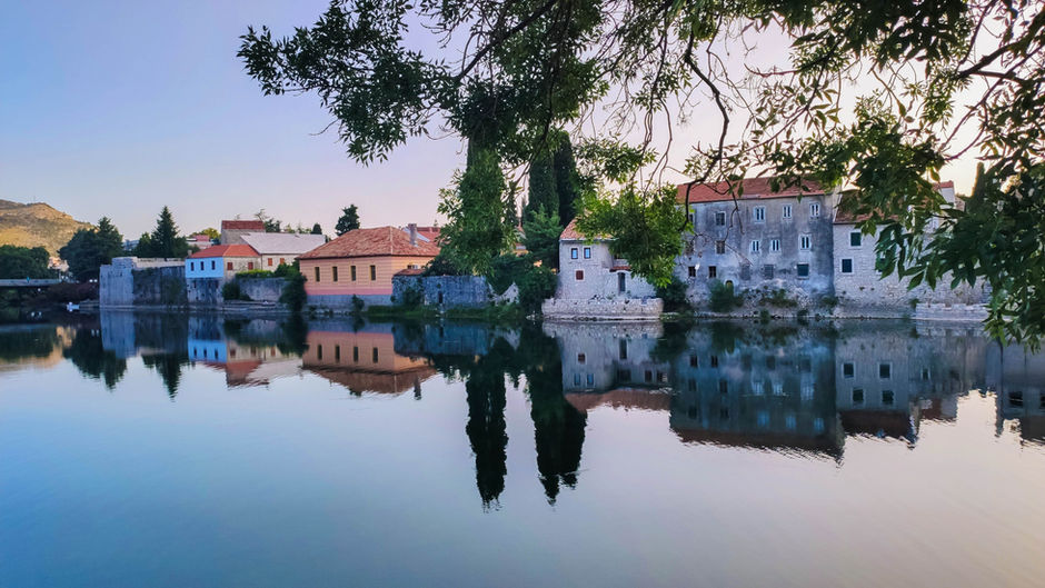 Tranquil riverside village with stone houses and tree branches reflected in still water at sunset.