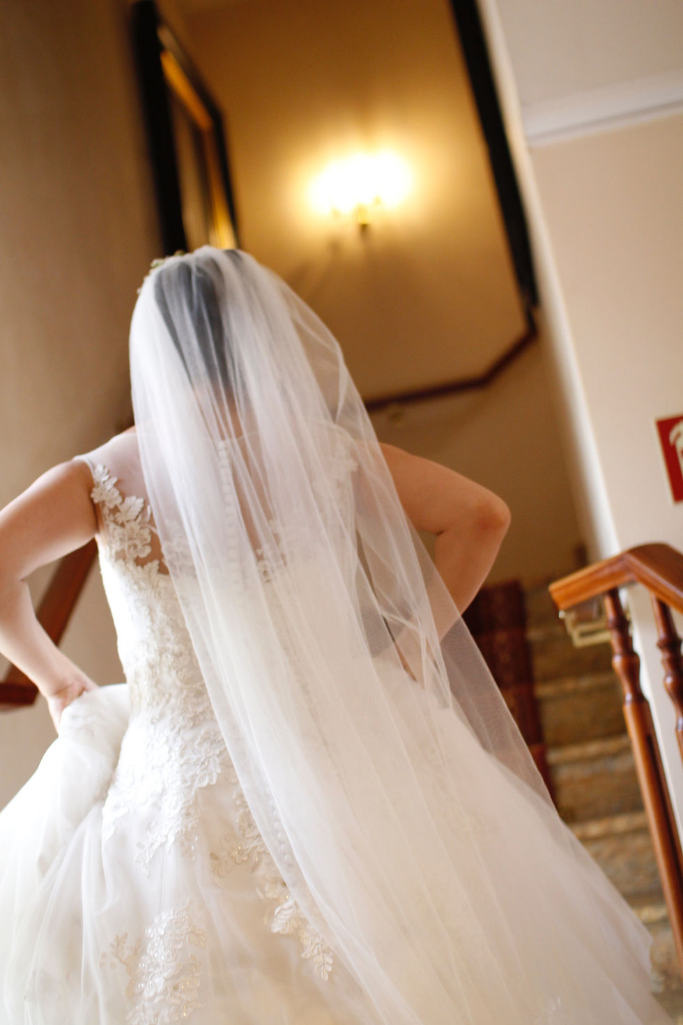 A bride walking up stairs. Her veil extends out of frame and has straight sides as it falls to the floor.